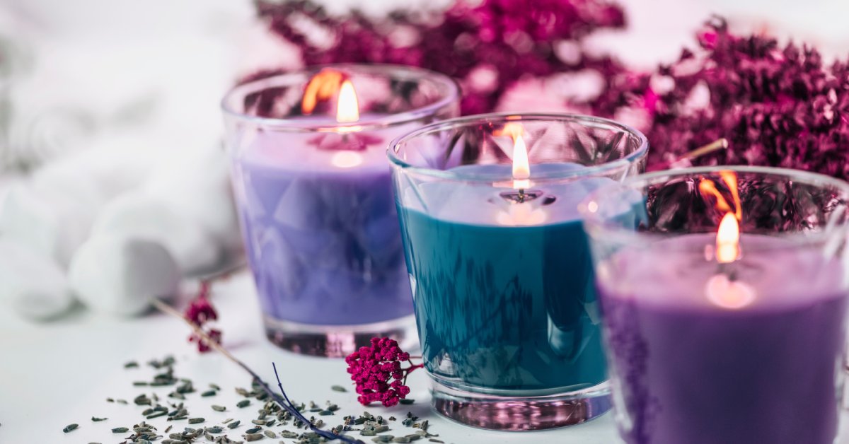 Three lit purple and blue candles in glass jars surrounded by dried lavender and pink flowers.