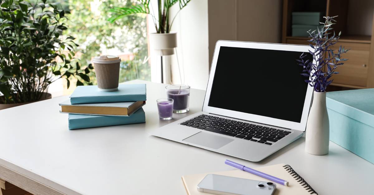 A white desk in an office with a laptop, blue box, plant, purple candles, notebook and pen, and books on it.