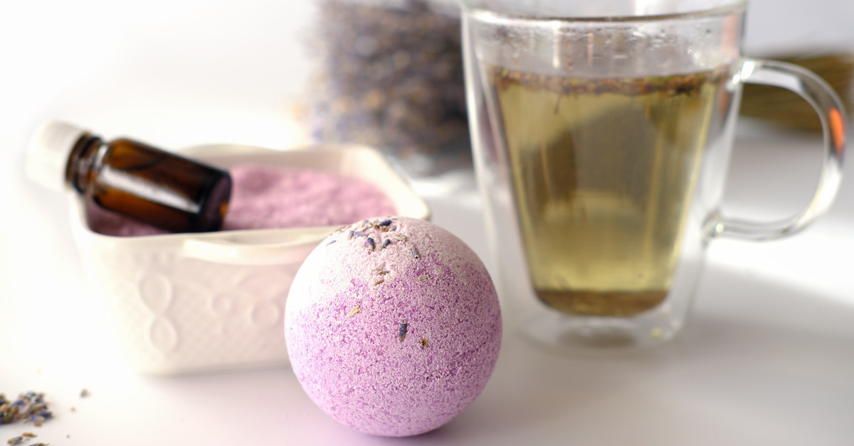 A lavender bathbomb next to a clear mug of tea. Behind the bathbomb and tea are lavender flowers.